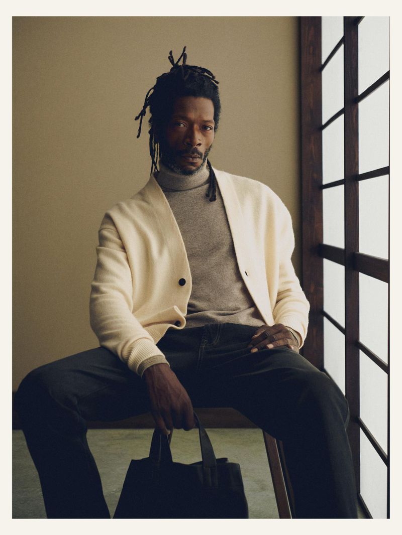 Black man sits on a stool in front of a decorative window. He is wearing a sweater and a cardigan and holding a black tote bag.