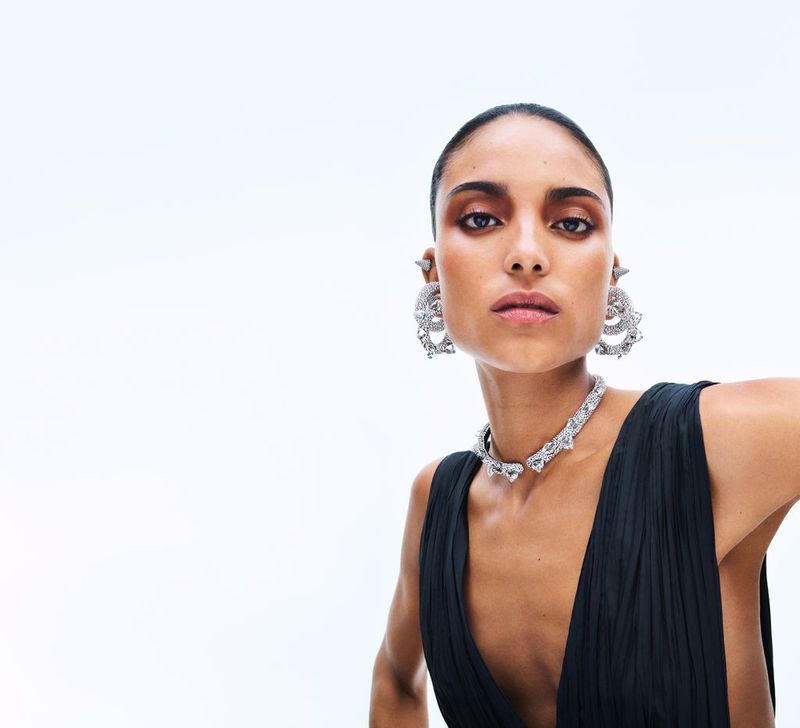 Close-up shot of a woman with short dark hair wearing jewelry and a black dress against a white background in a fashion setting.