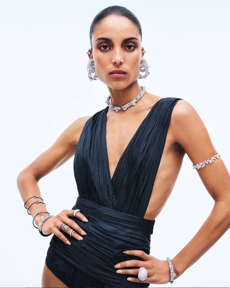 Fashion shot of a female model wearing a deep-V, dark blue leotard and a set of diamond jewelry against a bright white background.