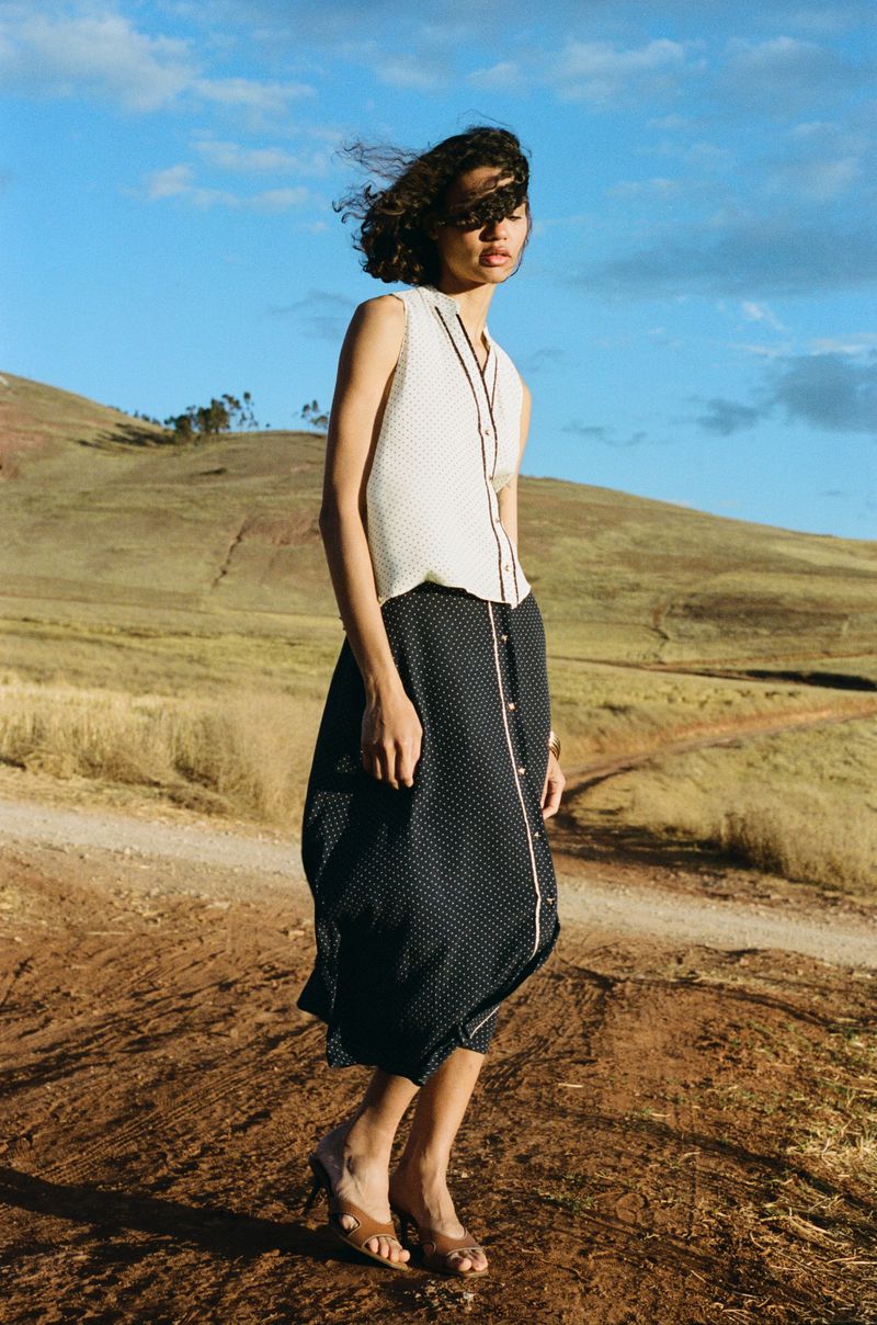 A young woman in a sleeveless top and midi skirt poses on a path in a rural landscape under a blue sky with scattered clouds.