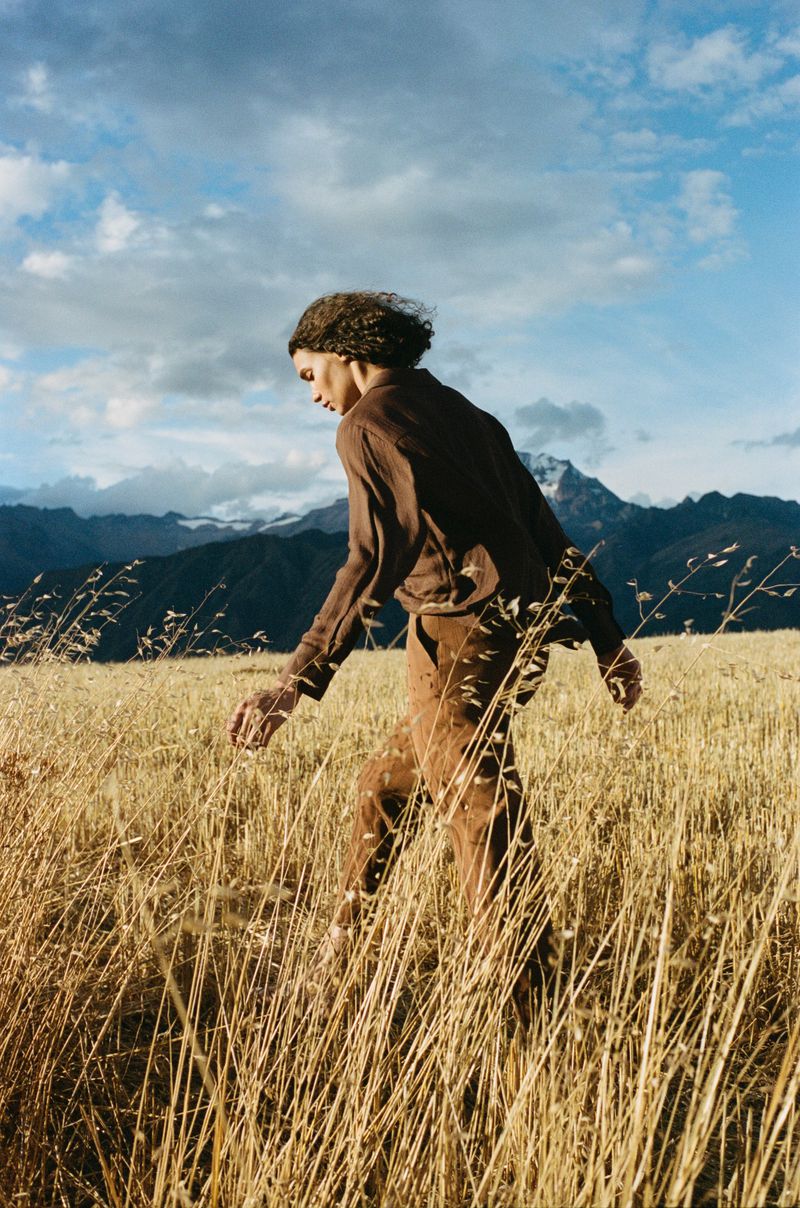 Woman walking through a golden grassy field with mountains and cloudy blue sky in the background; medium shot, eye-level angle, daytime.