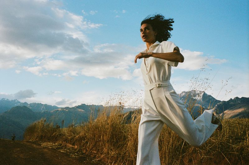 Model wearing a white jumpsuit running through a field with mountains under a cloudy sky.