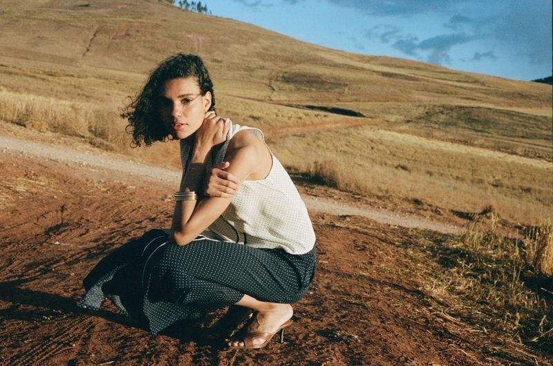 Woman in sleeveless top and patterned trousers squats on a hill, looking at the camera with a rural landscape under a blue sky.
