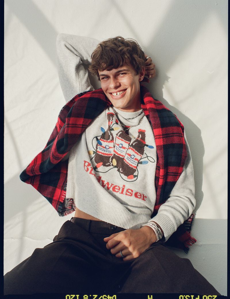 Smiling young man in a Budweiser sweater and plaid scarf, relaxing indoors with natural lighting and dark trousers, creating a casual fashion portrait.