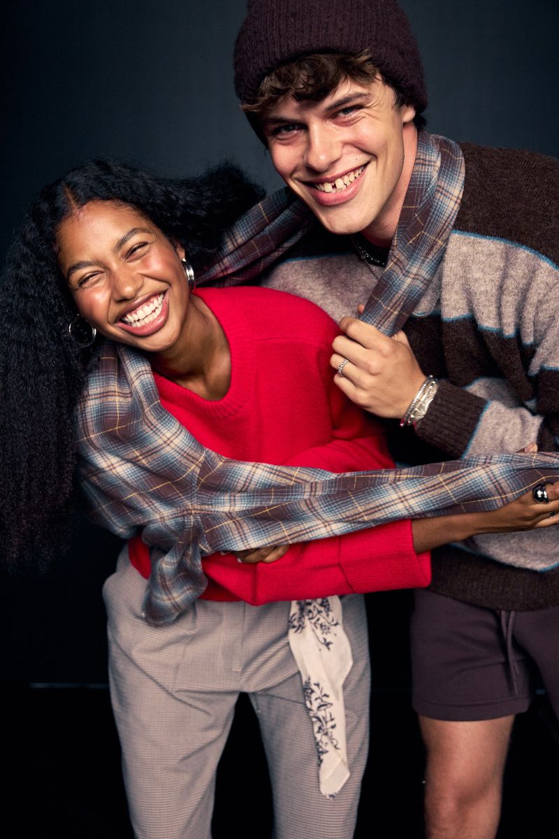 A smiling couple poses in a studio setting. The woman wears a red top and the man wears a striped sweater with a beanie.