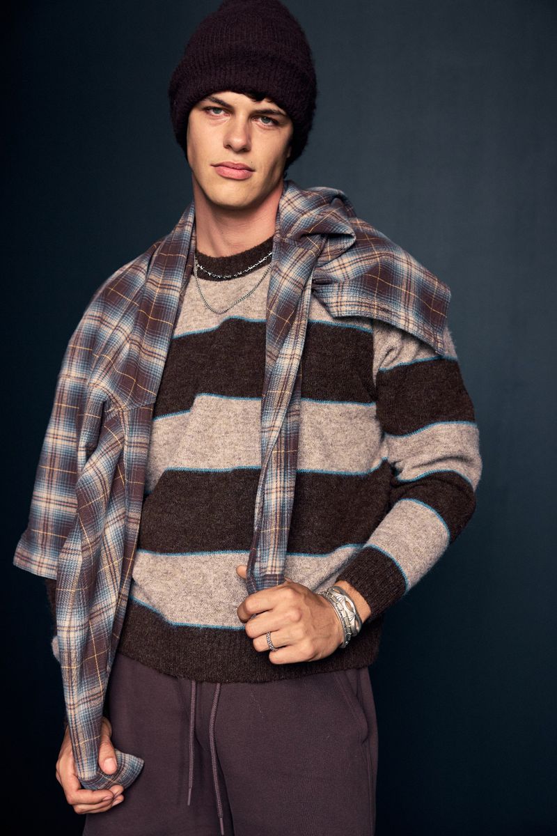 Male model in a brown striped sweater, beanie, and sweatpants poses in front of a dark background, wearing a plaid scarf.