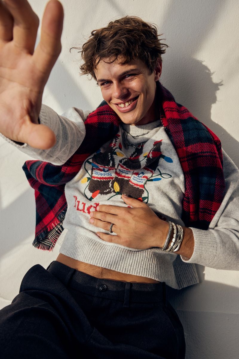 Portrait of a young, cheerful male model wearing a Budweiser-themed sweater and red plaid scarf, reaching toward the camera, indoors.