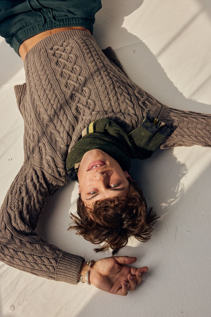 Young man lying on a white surface wearing a brown cable-knit sweater and green scarf, looking at the camera with a smile.