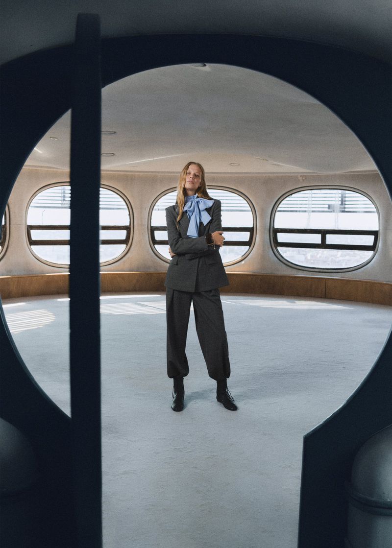A fashionable woman with blonde hair stands in a grey suit and blue bow in a room with oval windows, framed by a dark circular doorway.