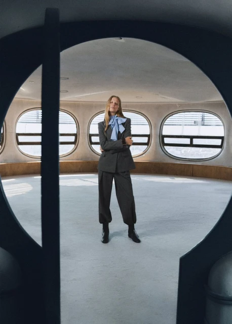 A fashionable woman with blonde hair stands in a grey suit and blue bow in a room with oval windows, framed by a dark circular doorway.