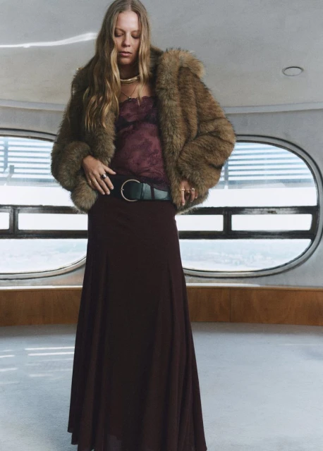 A woman in a fur coat and long skirt poses in front of a wall with round windows, showcasing a blend of fashion and architectural style.
