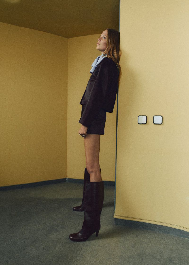 Indoor fashion shot: a woman in a brown suit and boots poses in a yellow room with blue carpet, looking up and away.
