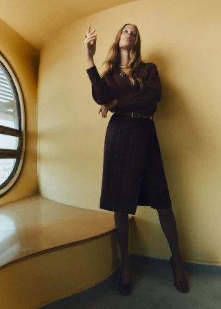 Woman poses in a plum-colored skirt suit near a curved window with yellow wall, wearing stockings and pumps in a modern indoor space.