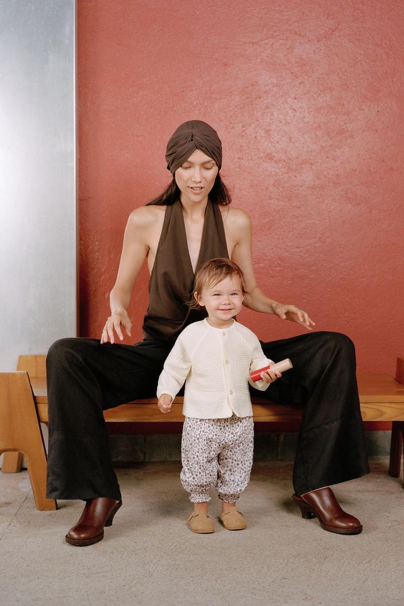 Mother and child together indoors: woman with turban sitting on a bench, toddler standing in front of her holding a small object.