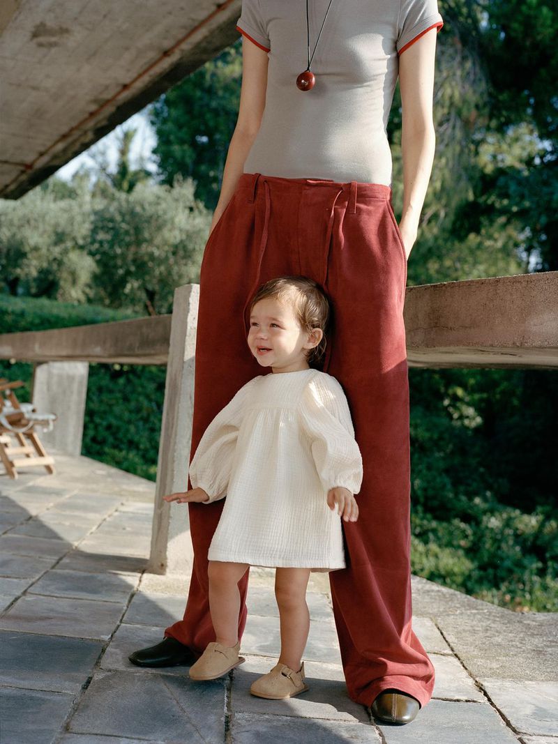 A young child in a white dress stands between the legs of an adult wearing wide-leg rust-colored pants, posing outdoors on a stone patio.