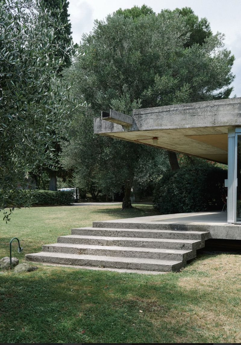 Exterior daytime view of a modernist building with concrete steps leading to the entrance, surrounded by trees and green grass.