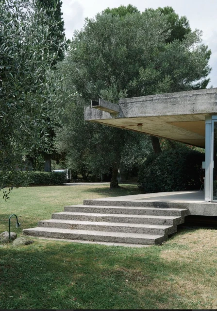 Exterior daytime view of a modernist building with concrete steps leading to the entrance, surrounded by trees and green grass.