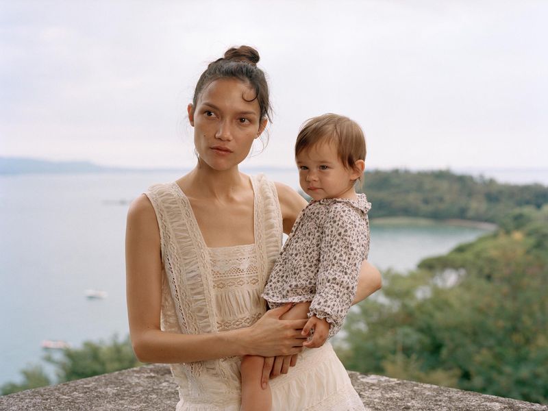 Mother holds her child in an outdoor setting with a body of water and trees in the background, both wearing light colored clothing.