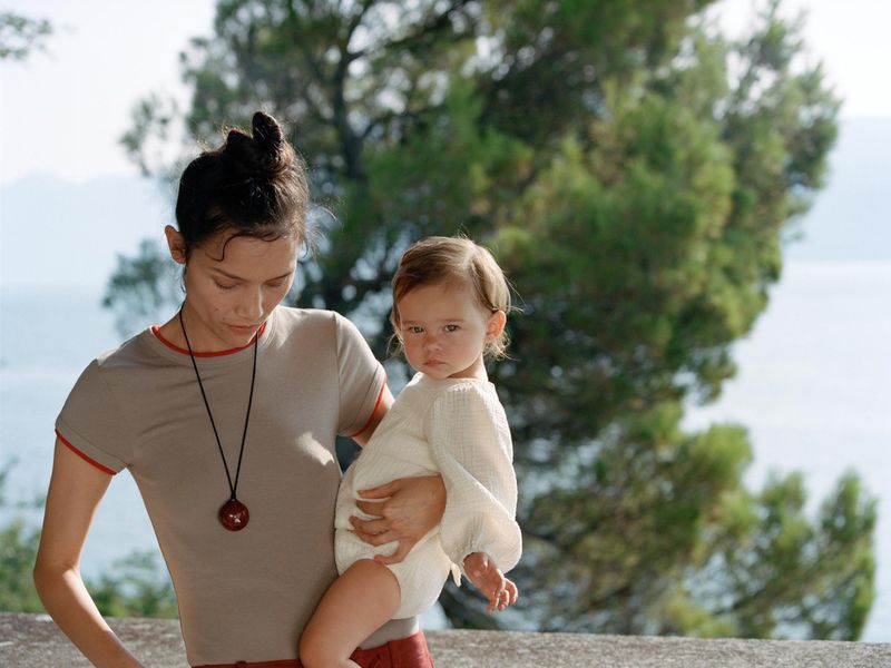 Woman in grey tee holds a baby in an outdoor setting with greenery. Both look at the camera. Sunny day with a view of the water.