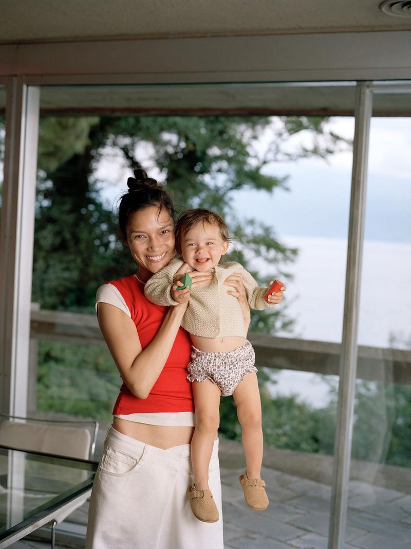 Happy mother holding her smiling baby daughter in front of a window with lake and greenery views, both looking at the camera.