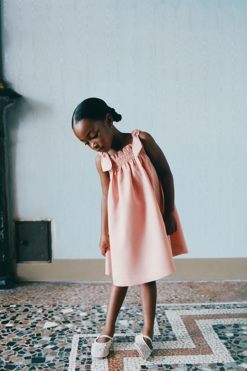 Adorable African-American child model in a light pink dress, posing indoors on a patterned tile floor, looking down in a children's fashion shoot.