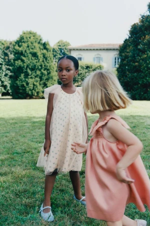 Two young girls pose outdoors in dresses, one with dark hair and fair dress, one with blonde hair and coral dress, grassy lawn.
