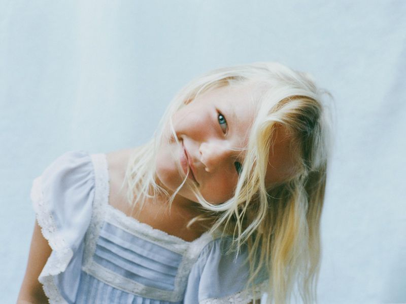 Close-up portrait of a fair-skinned young girl with blond hair smiling while tilting her head in a blue dress against a light background.