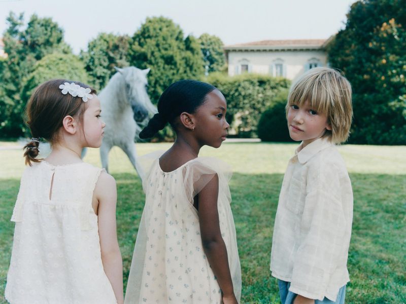 Three children stand in a green field with a white horse in the background near a house; the children are dressed in light-colored clothing.