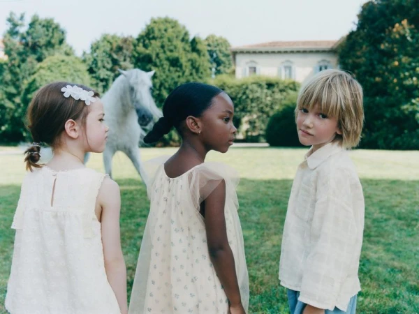 Three children stand in a green field with a white horse in the background near a house; the children are dressed in light-colored clothing.