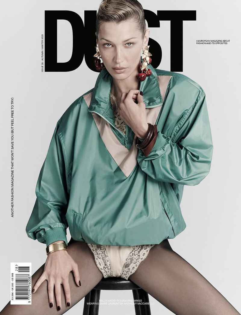 Bella Hadid poses on stool for DUST Magazine cover; she wears green jacket, bangles, and cherry earrings against a muted gray background.