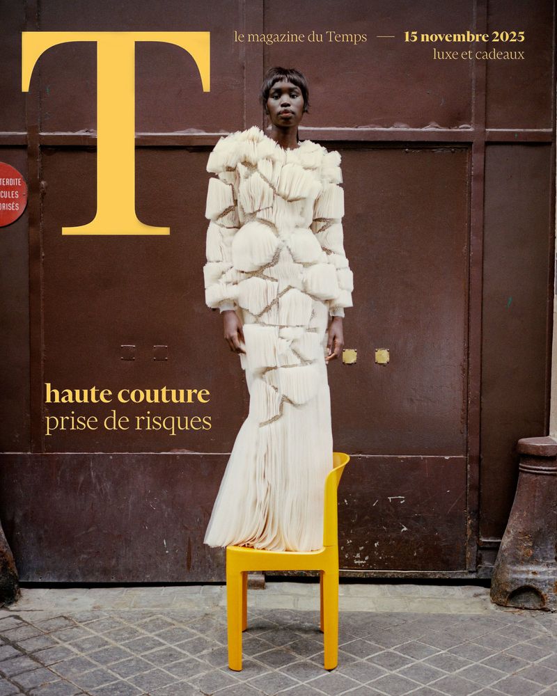 Magazine cover featuring a Black female model wearing a white haute couture dress, standing on a yellow chair against a brown door.