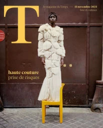 Model standing on a yellow chair wearing a structured white textured dress against a dark door.