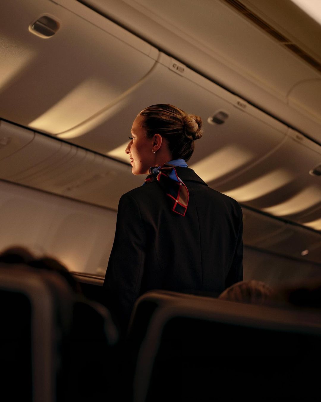 Flight attendant in dark blue uniform with patterned scarf stands in an airplane aisle with seats, overhead bins, and interior lighting.