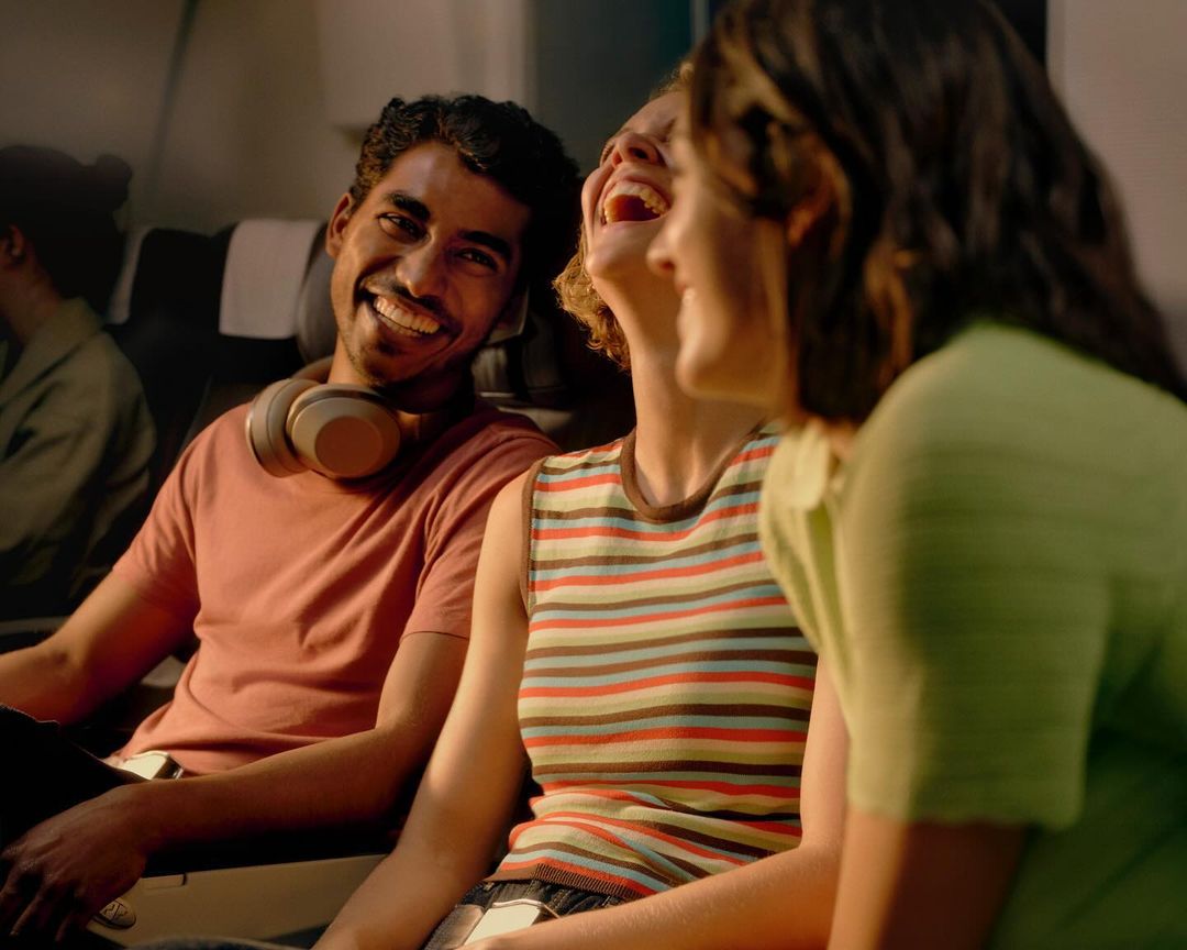 Three friends sit in adjacent airplane seats, talking and laughing. One is male, and the others are female. The man wears headphones.