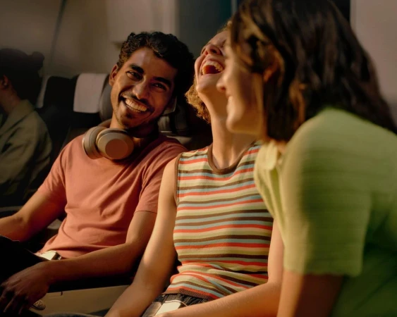 Three friends sit in adjacent airplane seats, talking and laughing. One is male, and the others are female. The man wears headphones.