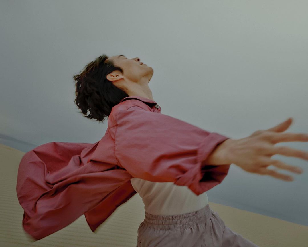 Young man posing outdoors in loose, pink outfit with outstretched arms.  Clear sky backdrop, low angle, fashion style.  