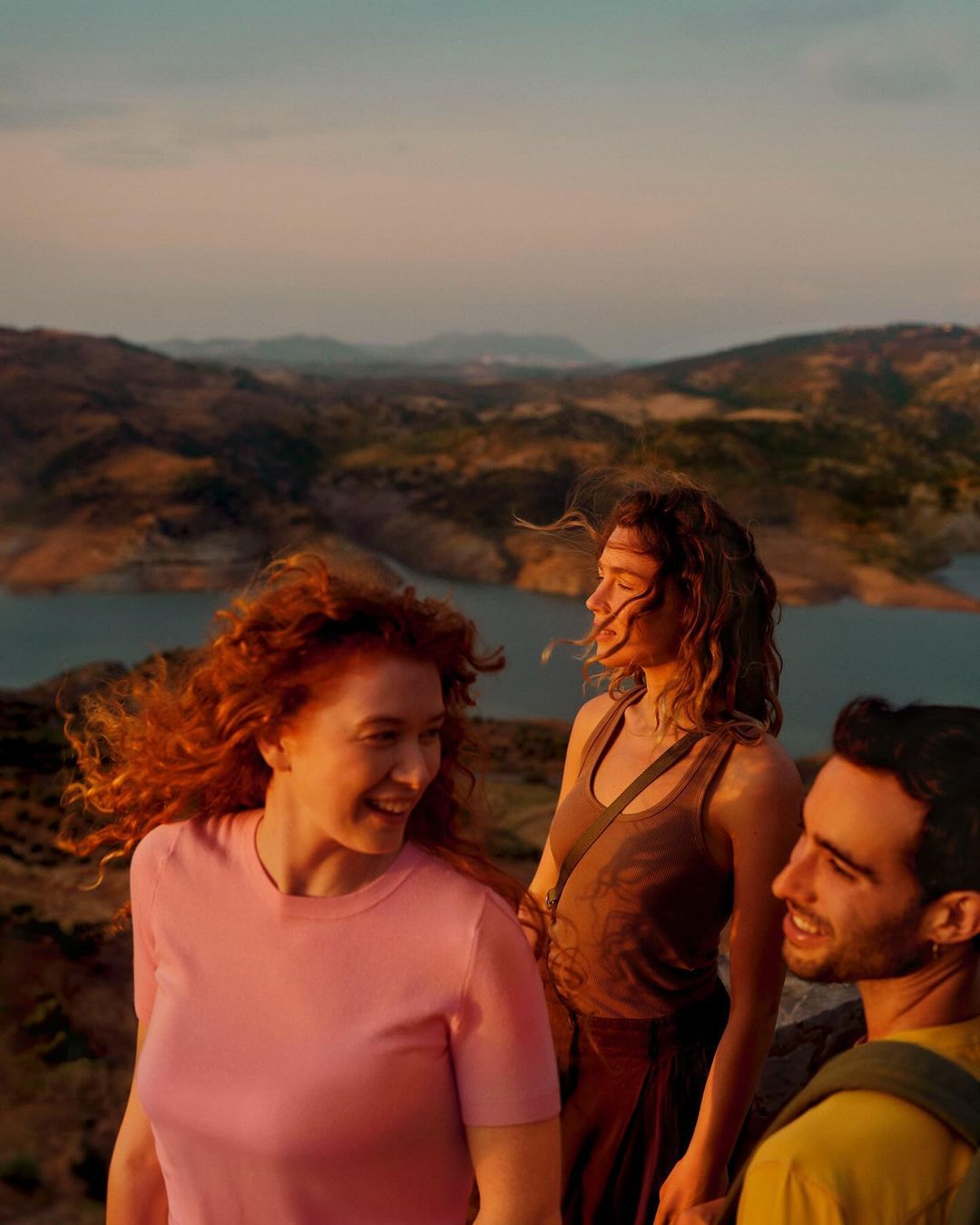 Three friends enjoy a scenic sunset view from a mountain top, featuring a red-haired woman smiling in the foreground and a lake in the background.