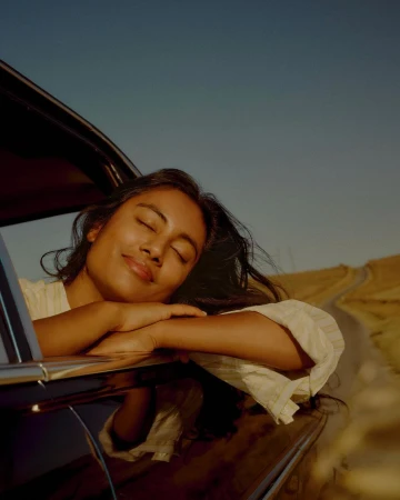 Woman relaxes in a vintage car, enjoying the roadside view during a golden hour road trip under a clear blue sky, creating a serene travel moment.