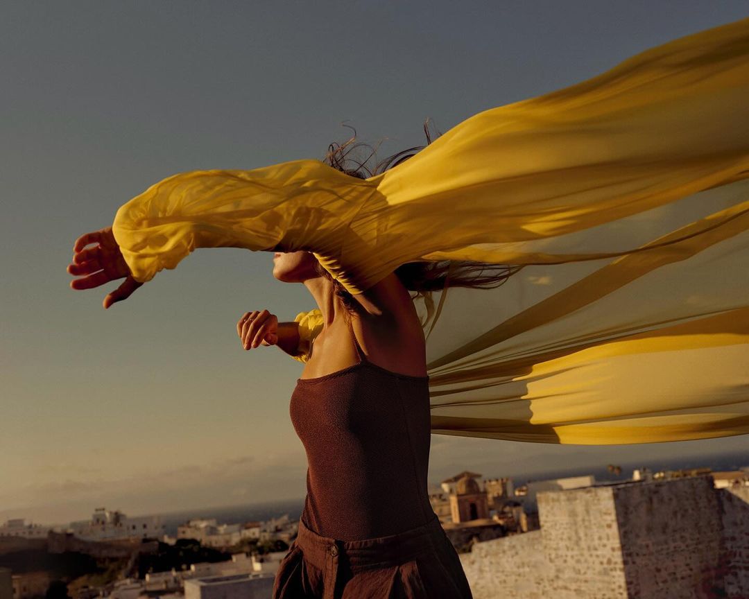 Woman stands on rooftop, a yellow fabric billowing in the wind. Background shows a whitewashed village under a clear blue sky at dusk.