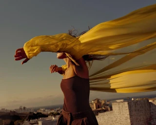 Model in yellow sheer fabric and dark brown top outdoors with town rooftops visible in the distance