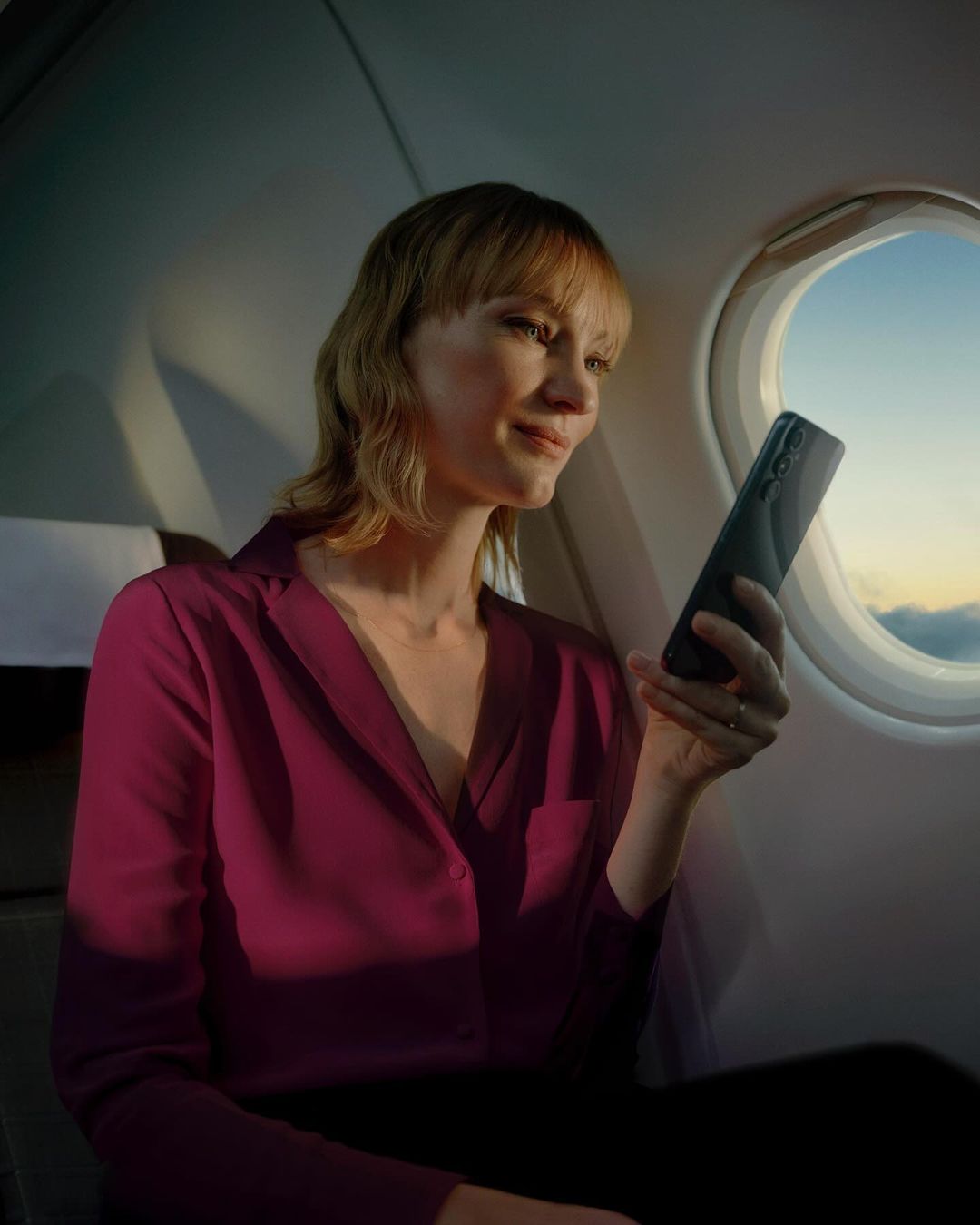 Adult woman using a black smartphone inside an airplane cabin at sunset. She's wearing a magenta blouse and looking at the screen.
