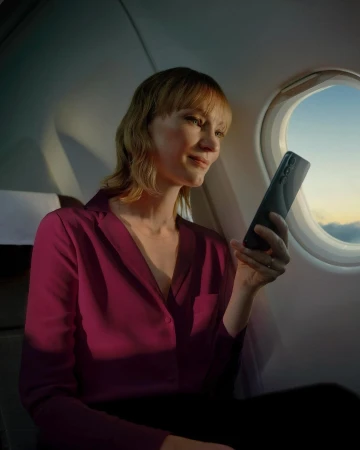 Adult woman using a black smartphone inside an airplane cabin at sunset. She's wearing a magenta blouse and looking at the screen.