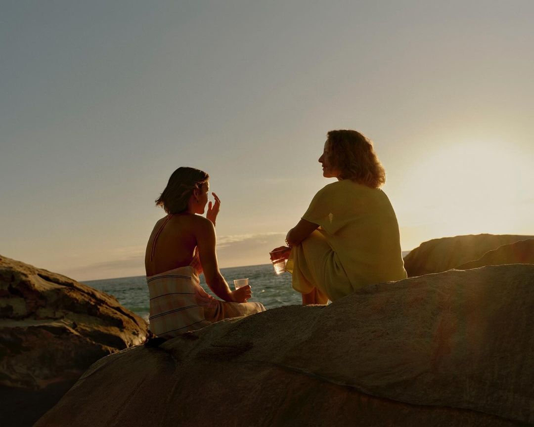 Two women sitting on coastal rocks at sunset holding drinks with the ocean in the background