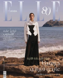 Model standing on a rocky shore wearing a structured white jacket and black skirt for Elle Arabia February 2025 cover.