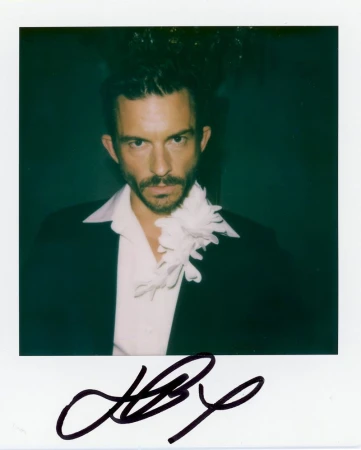 Polaroid portrait of an adult male with dark hair and a beard wearing a suit and tie with an off-white floral ruffle.