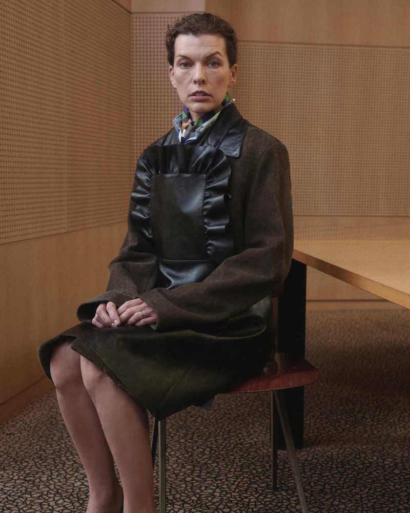 Woman in vintage-inspired fashion sits indoors, wearing a dark green coat and apron-like dress, presenting a blend of classic and modern style.