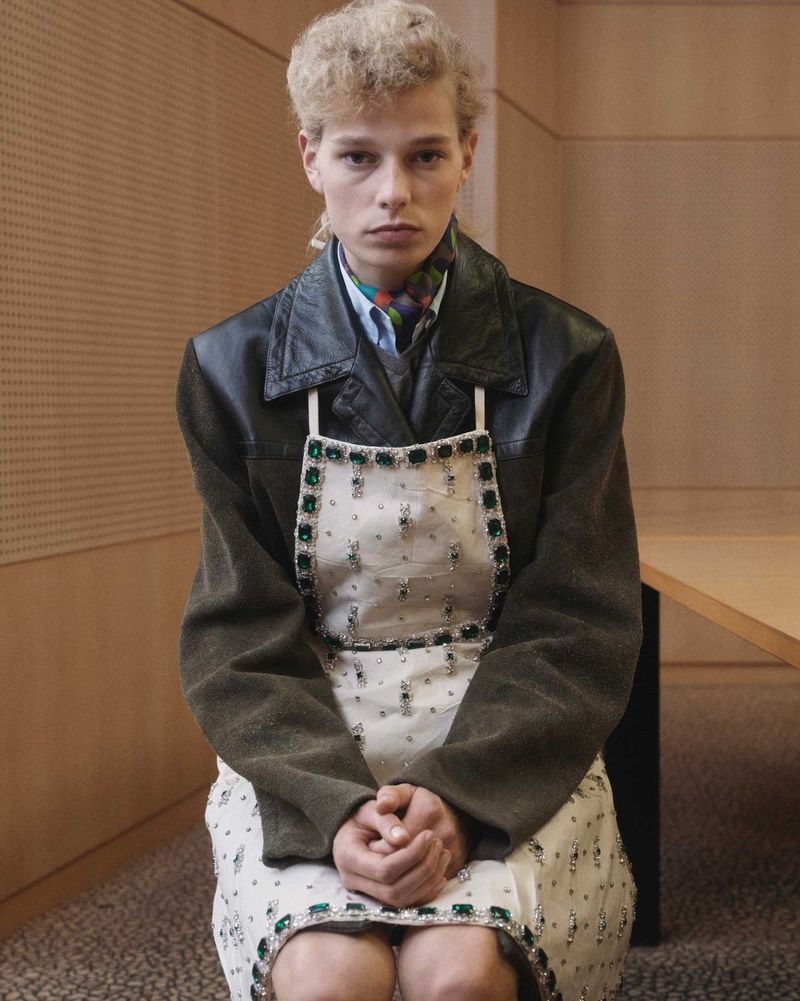Young adult sits indoors wearing a leather jacket, bib-front dress, and scarf, posing in front of a paneled wall.