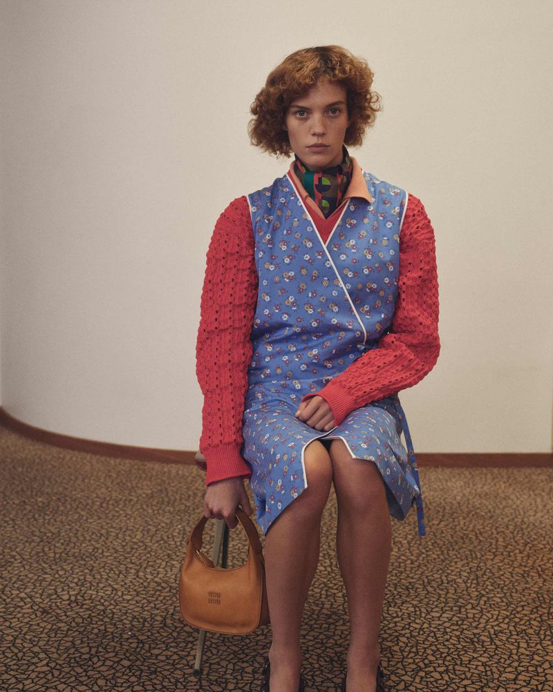 Indoor studio portrait of a fair-skinned adult female with auburn curls wearing a blue floral dress and holding a tan handbag.