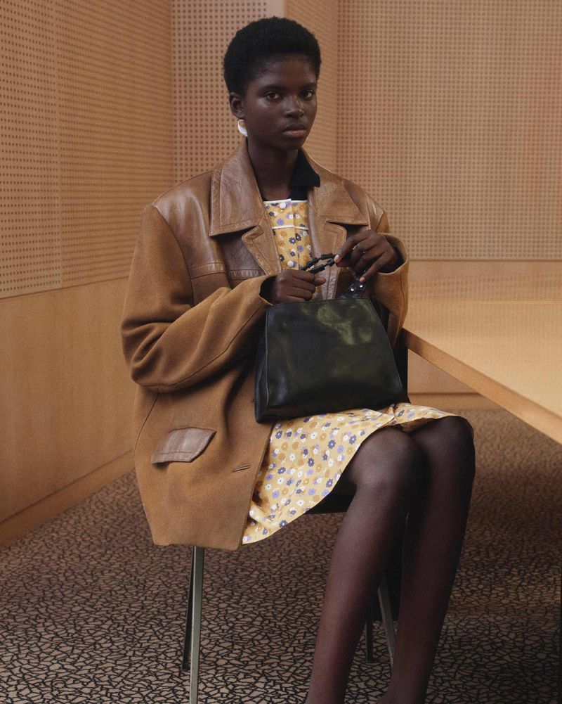 Model wearing a brown jacket and patterned dress holding a black handbag while seated in an office.