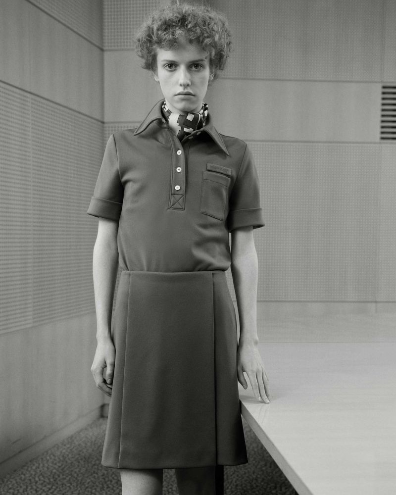 Monochrome portrait of a slender young adult with curly hair wearing a polo shirt and skirt, posing indoors against a textured wall.